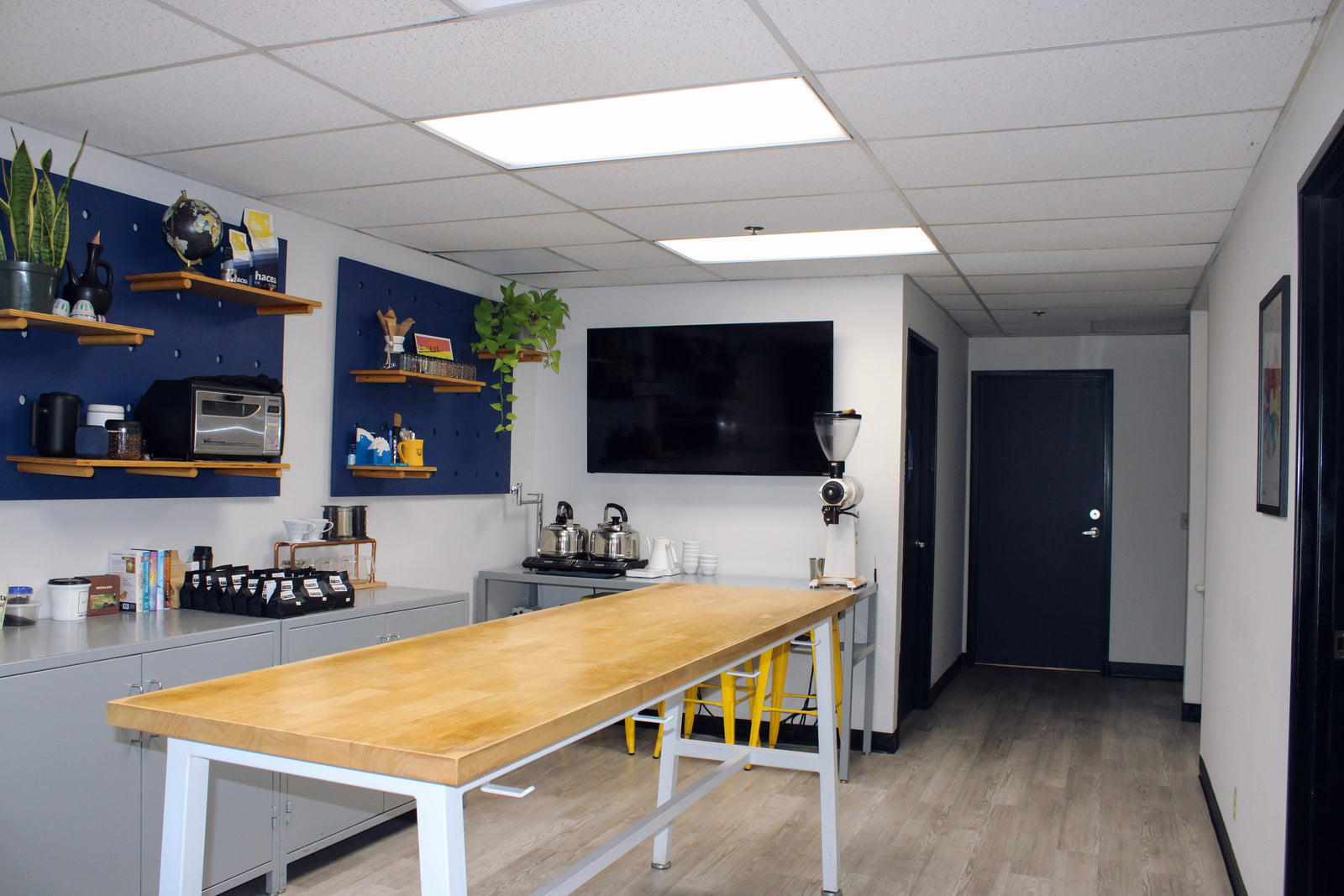 Hacea cupping lab with a long wooden-topped cupping table in the foreground, blue accent walls with shelves, coffee equipment, a mounted TV, and light wood flooring.