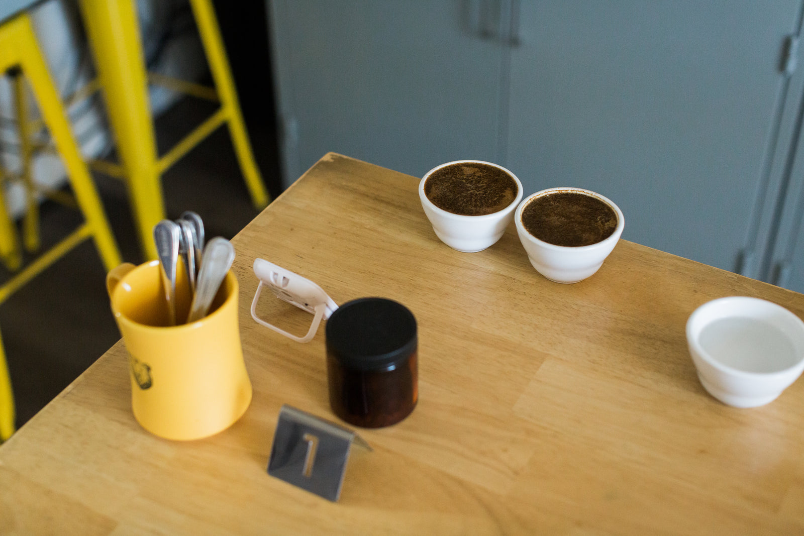 A wooden tabletop with a yellow mug holding utensils, a small amber bottle with a black lid, a white cup, and two small cupping bowls with steeping coffee.