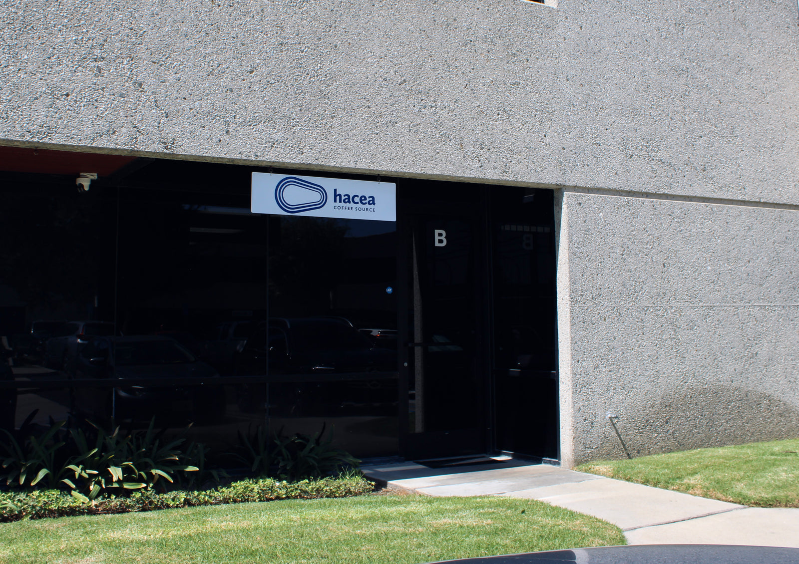 Glass windows and door on a concrete building with Hacea Coffee Source sign showing building location entrance.