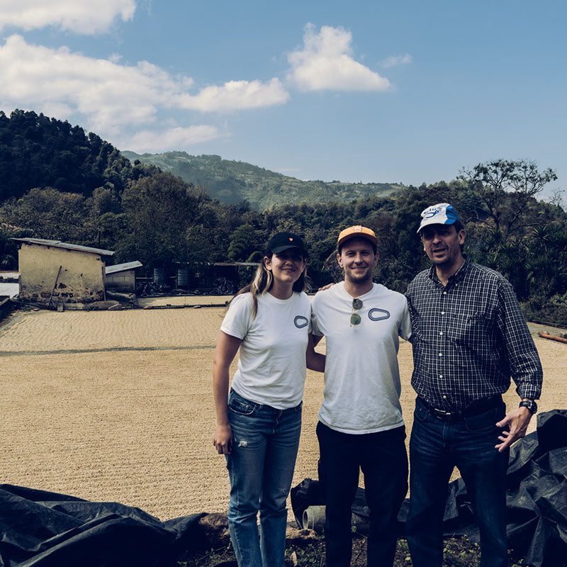 Three people posing in front of a patio of drying coffee in Guatemala.