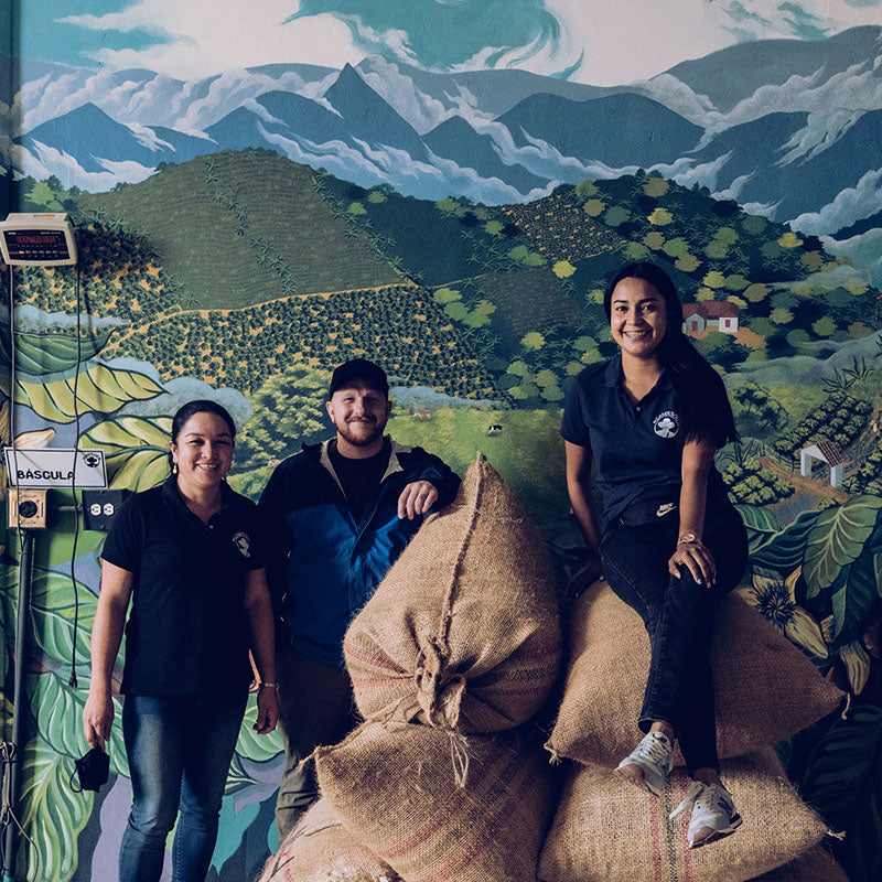 Three people posing in front of a painted mural around sacks of coffee in Colombia