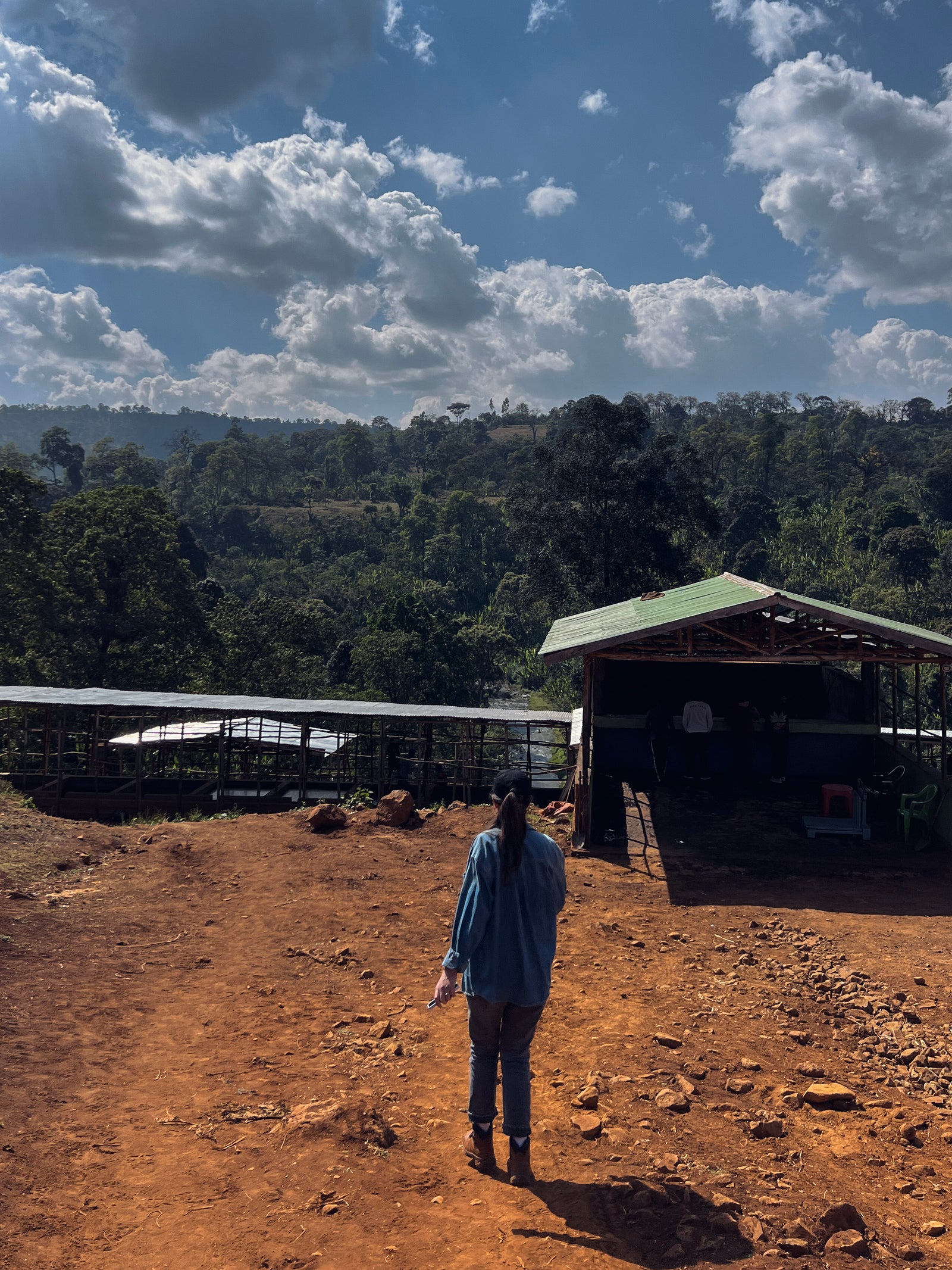 Hacea team member standing on a dirt path with a scenic view of the Bishan Wate washing station on a hill next to a river.