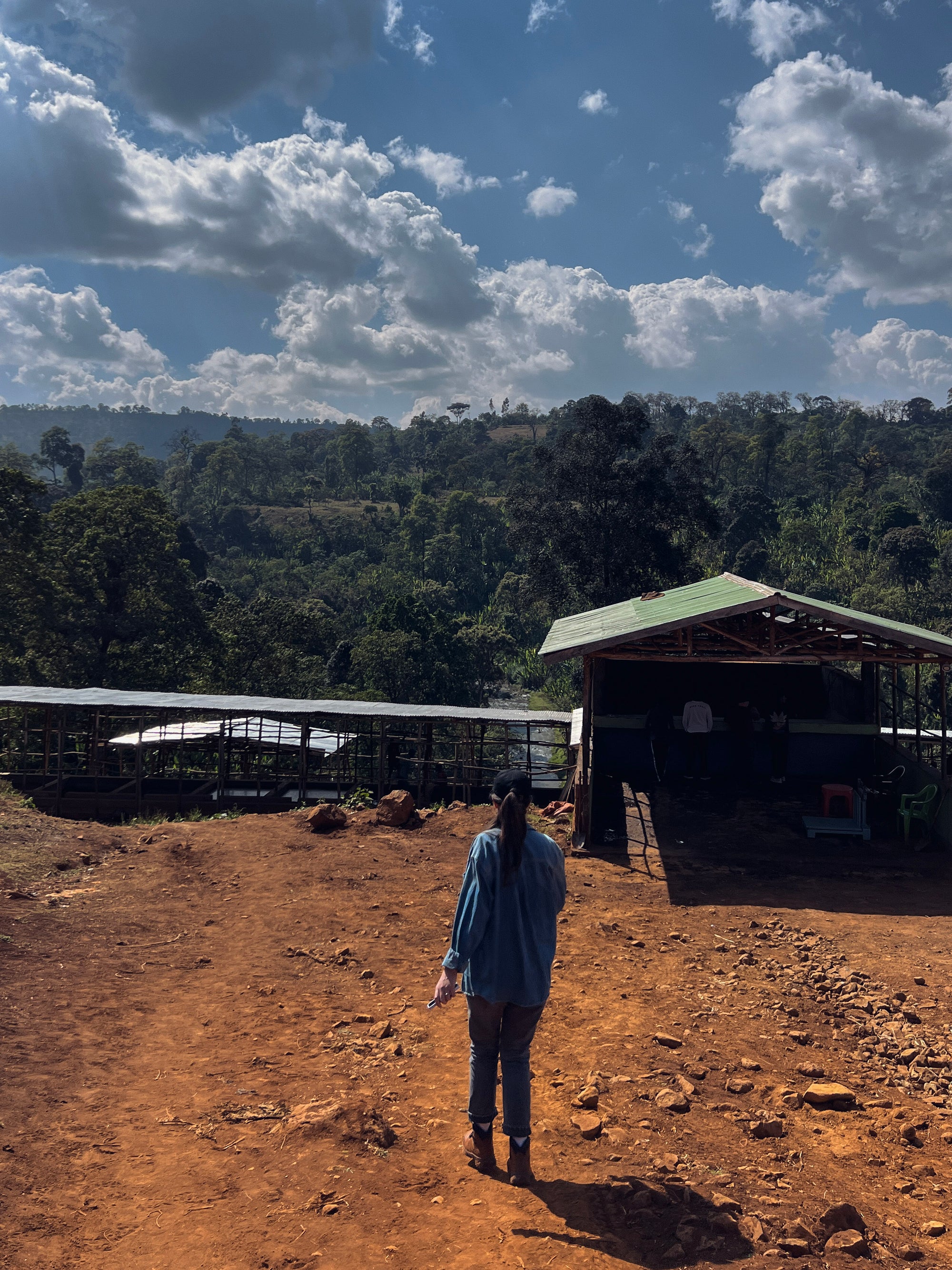Hacea team member standing on a dirt path with a scenic view of the Bishan Wate washing station on a hill next to a river.